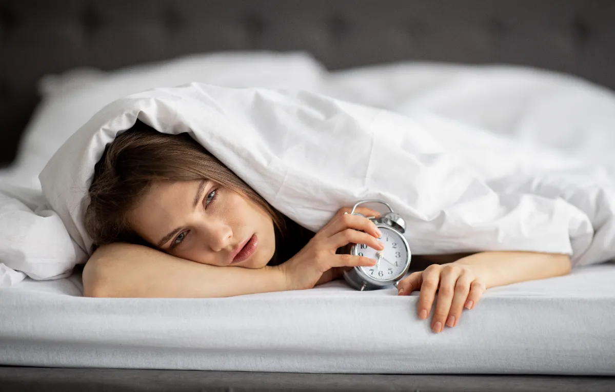 sleepy woman lying in bed holding alarm clock utc